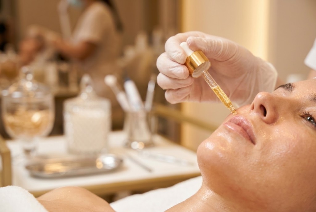 Esthetician's gloved hand applying a golden serum dropper to a client's glowing cheek during a clinical facial treatment